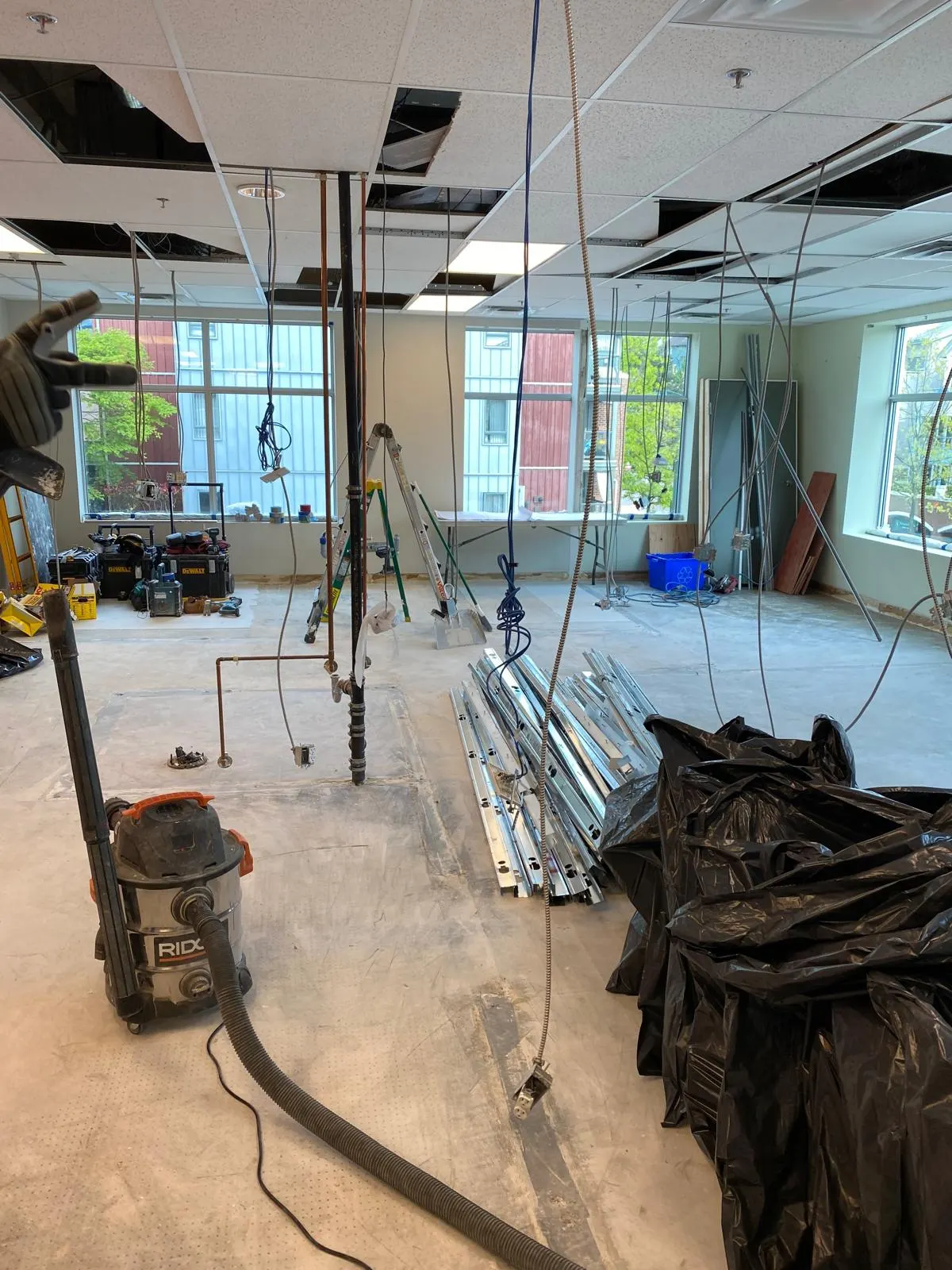 Open ceiling with demolished tiles and exposed rough-in plumbing, a wet vacuum staged on the floor, city buildings visible through the windows