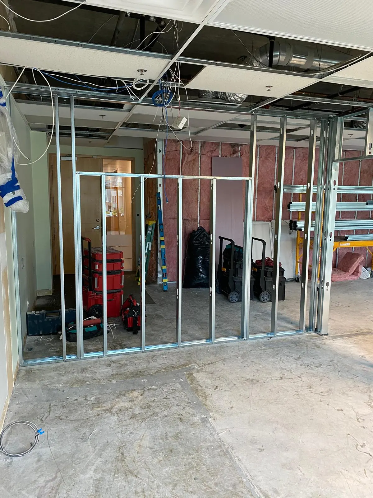 Steel-stud framing in progress beside an existing wood door, tool carts and insulation staged along the wall