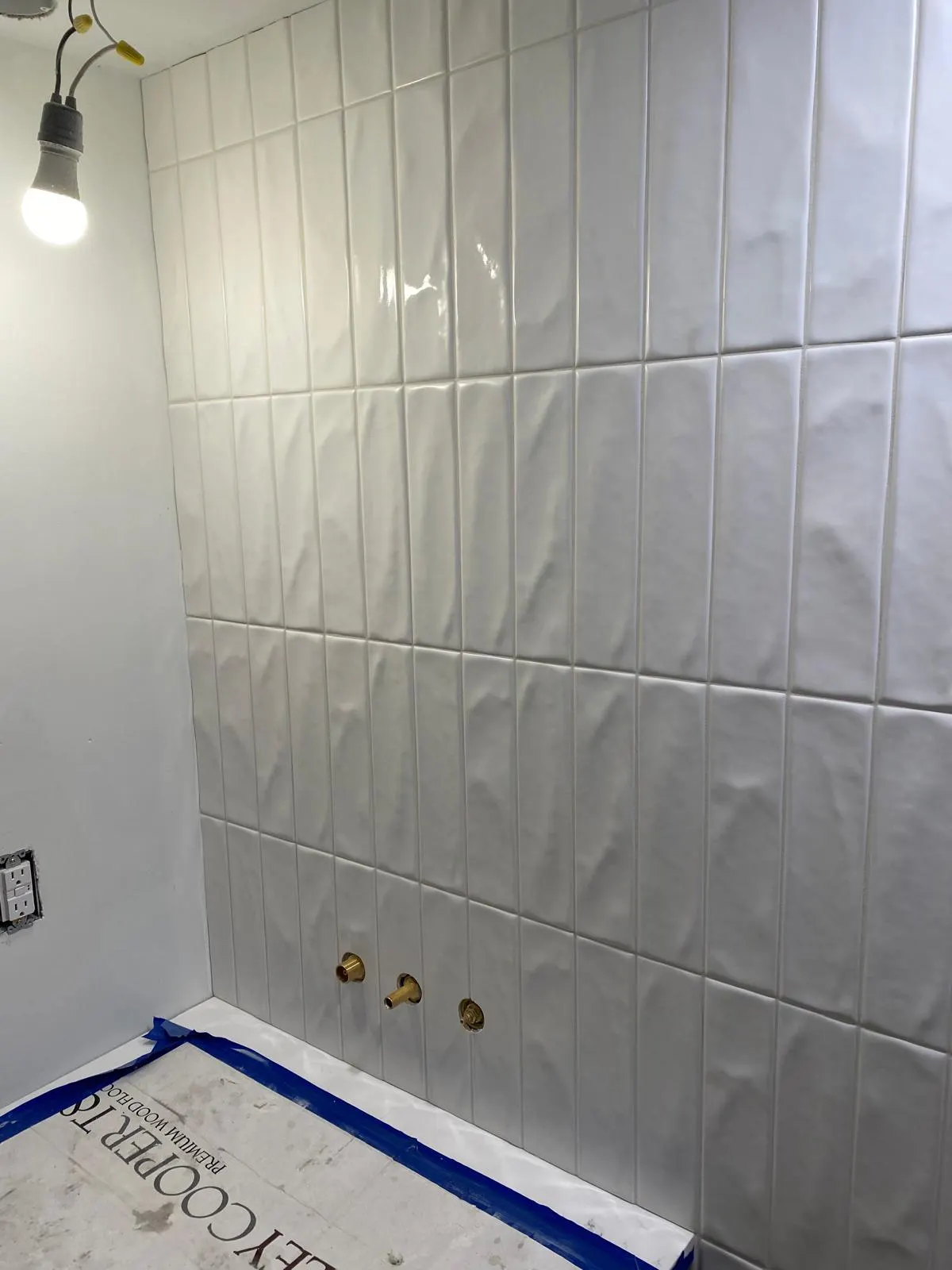 Bathroom shower mid-renovation with white subway tile and brass fixtures, protective tape still on the floor
