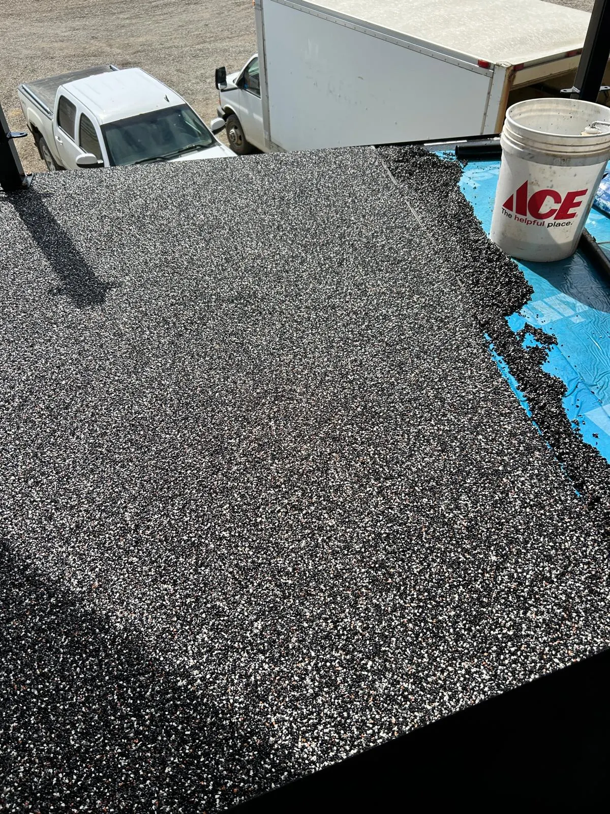 Close-up of a poured rubber surface being applied and rolled out on a flat exterior deck, ACE paint bucket in frame